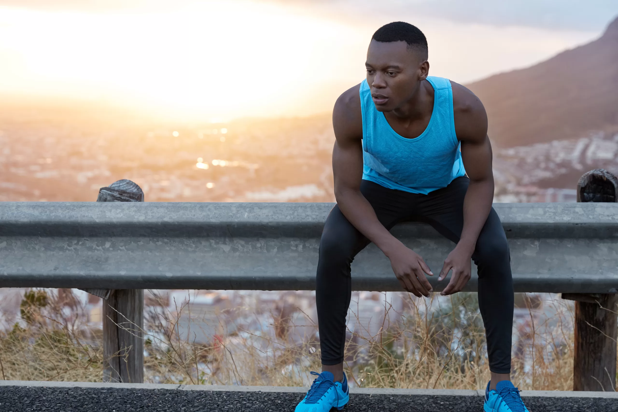 image of tired african american man with thoughtful expression, keeps gaze down, feels tired after intense training, sits at road sign, beautiful sunrise in background with copy space for information