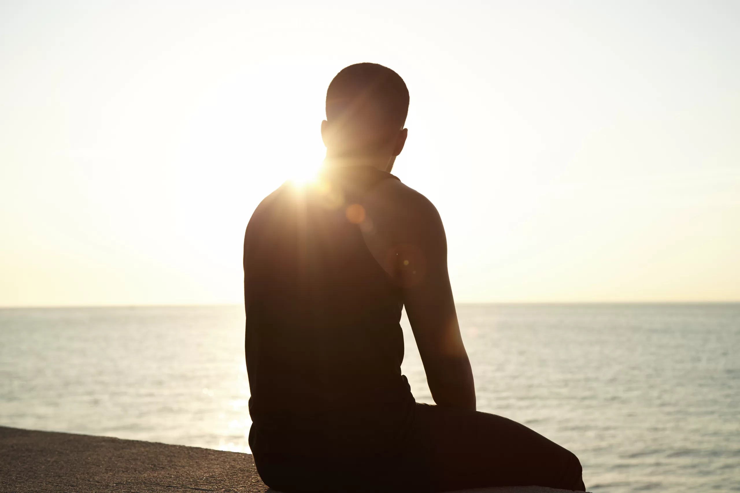 silhouette of pensive african young male sitting on sidewalks at seaside watching sun setting over sea after outdoor training, waiting for his girlfriend, thinking about his relationships. flare sun