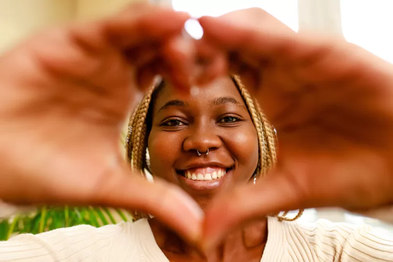 hispanic woman showing love with hands in heart shape expressing healthy and marriage symbol near window and green plant at home